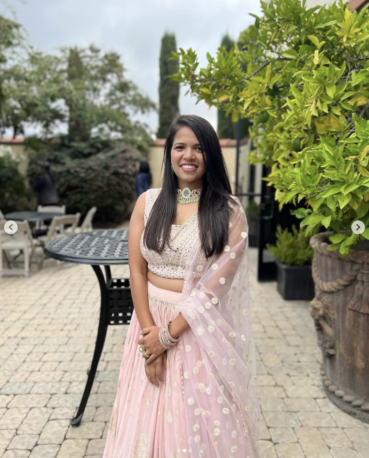 Bhavi smiling in a blush lehenga at an outdoor celebration.