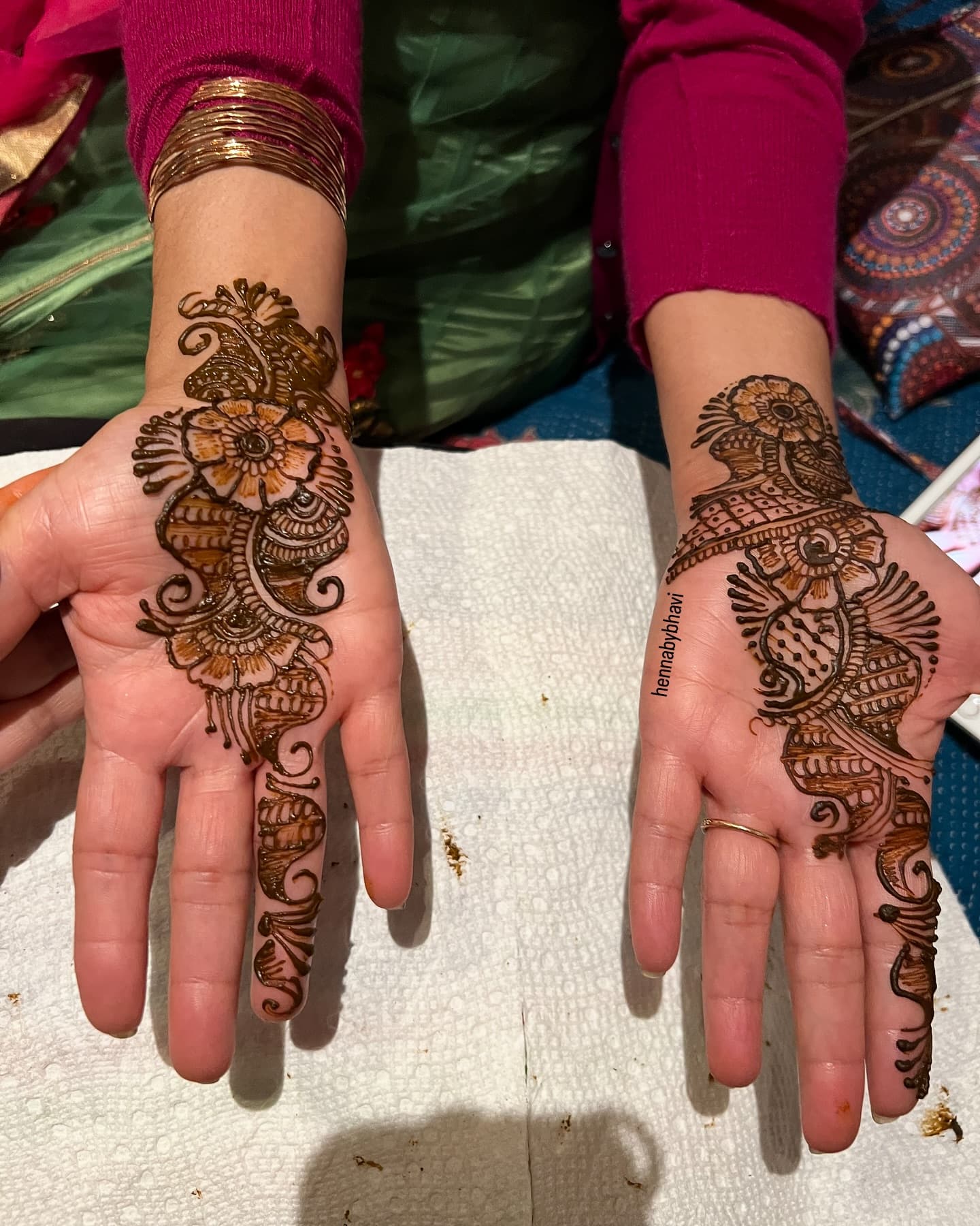 Two hands with floral paisley henna beside bangles and a maroon sleeve